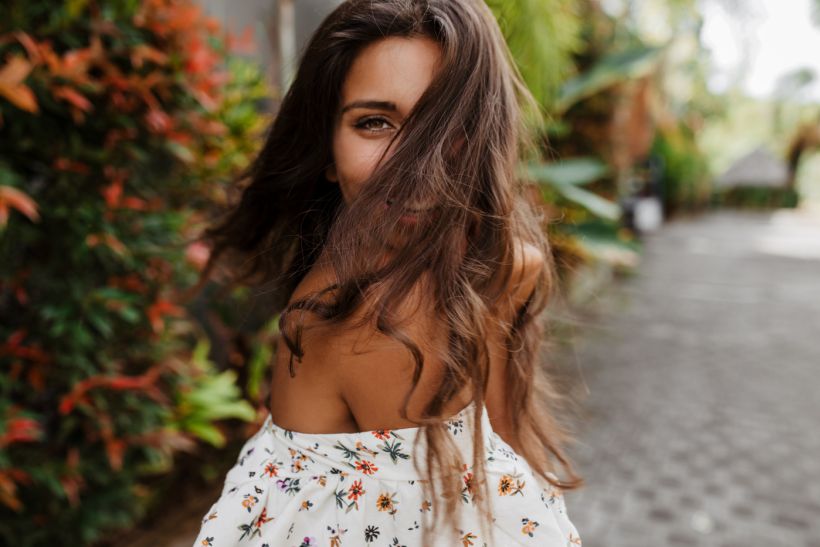 Charming tanned lady in white top with colorful flowers turns to camera and plays her hair against / envato